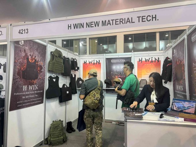 H Win's booth at a trade show, showcasing their range of bulletproof vests and helmets, with staff interacting with customers